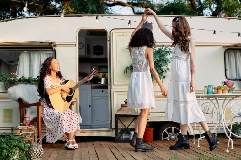 Young happy women have fun together playing guitar and dancing outdoors near their camper van during summer vacation