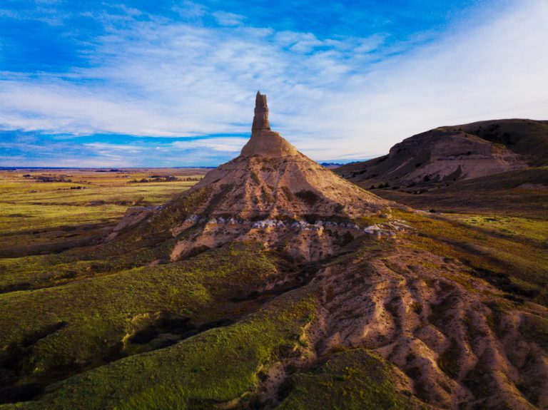 A tall, cone-shaped rock formation topped with a narrow rock tower sits next to a bluff and open fields.