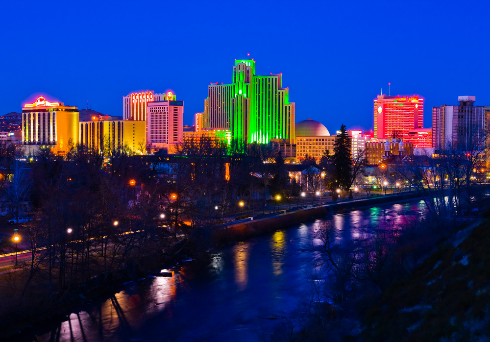 A cityscape at night. Green, red, and yellow lights shine on the buildings that stand beside a waterway.